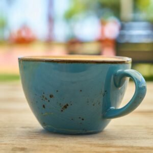A teal ceramic mug of coffee sits on a wooden table outdoors, showcasing a bokeh background.