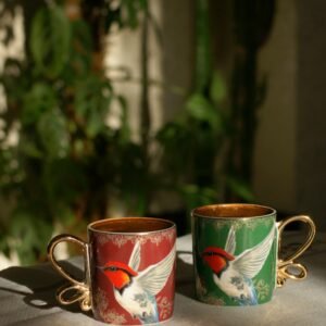 Two colorful ceramic mugs with bird designs placed on a sunlit table surrounded by indoor plants.