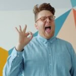 Smiling man in blue shirt making an okay sign against colorful wall.