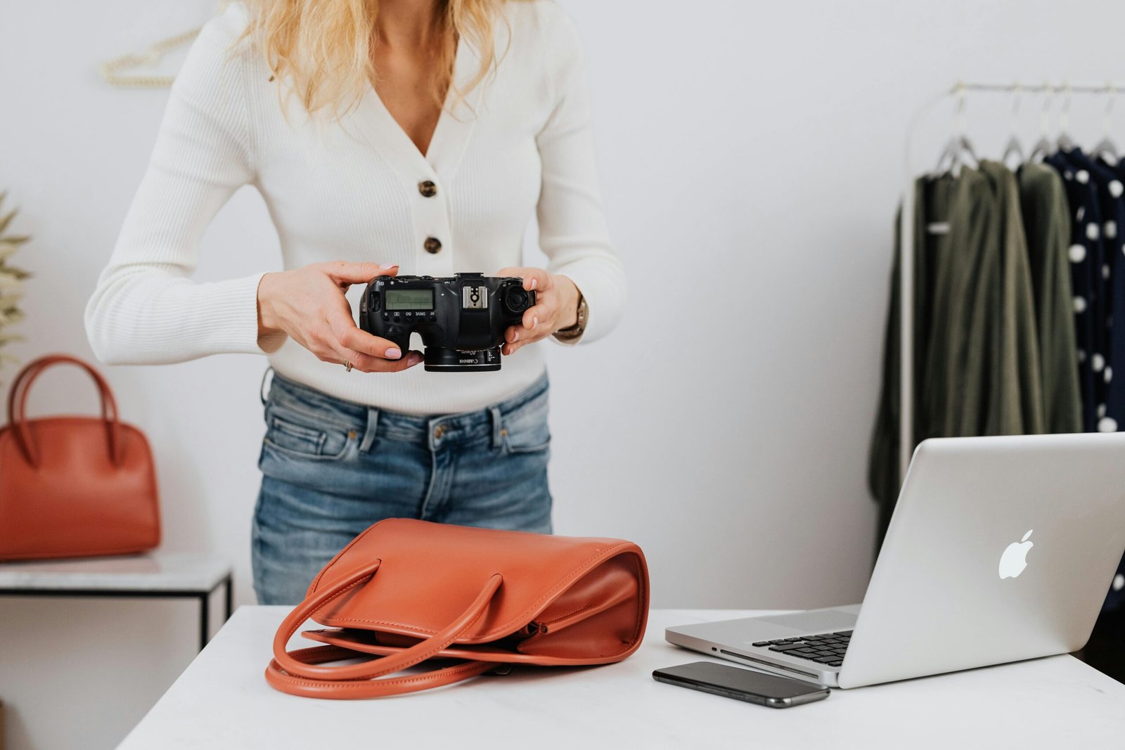 Woman photographing fashion items indoors with DSLR, handbag, and laptop.